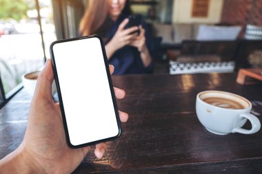 Kafede arka planda telefon kullanan bir kadınla birlikte boş bir masaüstü ekran ile siyah cep telefonu tutan bir elin görüntüsü.