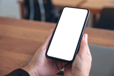 Siyah cep telefonlu, boş beyaz ekranlı ve ahşap masadaki laptoplu ellerin görüntüsü.
