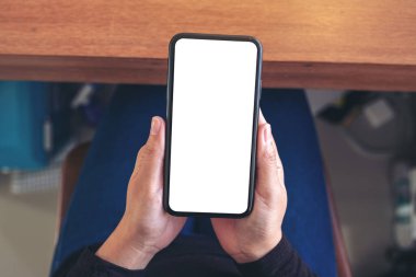 Üst görünümde, bir sandalyede otururken siyah cep telefonunu beyaz ekran ile tutan bir kadının ellerinin görüntüsü.