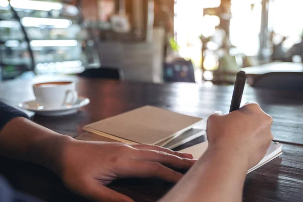 カフェのテーブルの上にコーヒーカップが置かれた空白のノートに書いている女性のイメージを閉じる — ストック写真