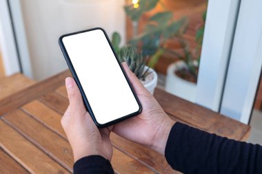 Ellerin ahşap masadaki boş beyaz ekranlı siyah cep telefonunu tutarken görüntüsü.