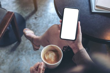 Kafede oturup kahve içerken, elinde beyaz ekranlı siyah cep telefonu olan bir kadının görüntüsü.