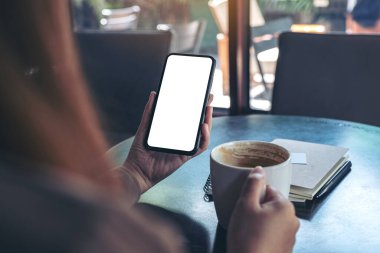 Kafede oturup kahve içerken elinde beyaz ekranlı siyah cep telefonu olan bir kadının görüntüsü.