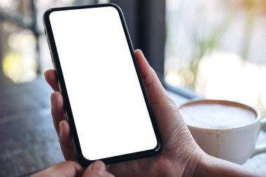 Kahve masasında kahve fincanı ve boş masaüstü ekranı olan siyah cep telefonunu tutan ellerin görüntüsü.