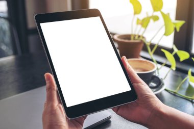 El ele tutuşmanın ve siyah tablet tableti kullanmanın resmi. Boş beyaz masaüstü ekran, defter, yeşil yapraklar ve ahşap masa üzerinde kahve fincanı.