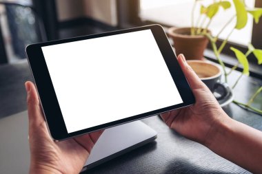 El ele tutuşmanın ve siyah tablet tableti kullanmanın resmi. Boş beyaz masaüstü ekran, defter, yeşil yapraklar ve ahşap masa üzerinde kahve fincanı.