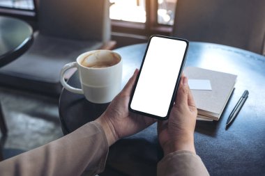 Ellerinin siyah bir cep telefonunu tutarken ve kullanırken çekilmiş bir resim. Boş ekranlı, defter ve kahve fincanı ile ahşap masa üzerinde izlemek için.