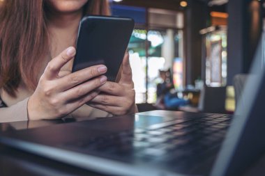 Kafedeki masada dizüstü bilgisayarı olan, elinde tutan, kullanan ve akıllı telefona bakan bir kadının yakın plan görüntüsü.