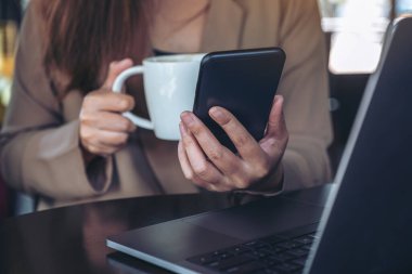 Kahve içerken ve kafede dizüstü bilgisayarla çalışırken elinde tutan, kullanan ve akıllı telefona bakan bir kadının yakın plan görüntüsü.