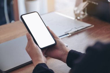 Siyah cep telefonlu, boş beyaz ekranlı ve ahşap masadaki laptoplu ellerin görüntüsü.