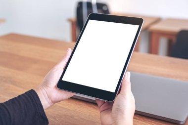 Ofisteki ahşap masadaki defter ile birlikte boş beyaz masaüstü ekranı olan siyah tablet bilgisayarın el ele tutuşunu ve kullanımını görüntüle