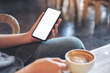 Modern kafede kahve içerken elinde beyaz cep telefonu olan bir kadının görüntüsü.