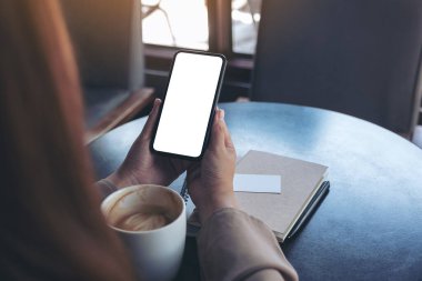 Siyah bir cep telefonu tutan ve kullanan bir kadının görüntüsü. Boş ekran ile ahşap masada defter ve kahve fincanı ile izlemek için.