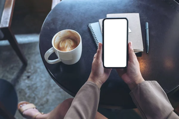 Top view mockup image of woman holding black mobile phone with blank white screen with notebooks and coffee cup on table