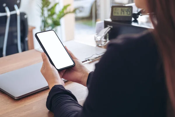 Mockup image of a woman holding black mobile phone with blank white screen with laptop on wooden table