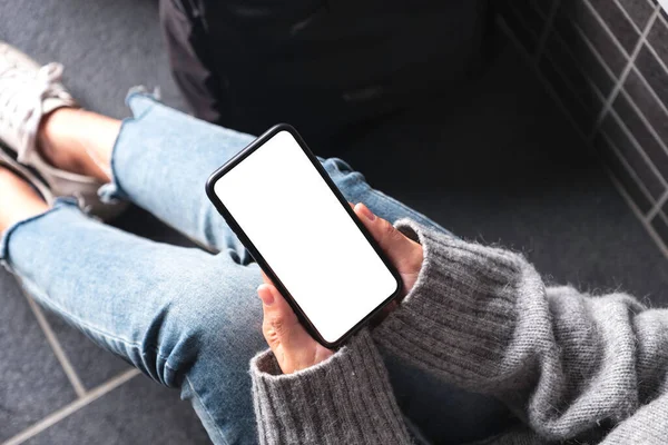 Üst görünümde, elinde boş masaüstü ekran ile siyah cep telefonu olan bir kadının yerde bavulla otururken görüntüsü var.