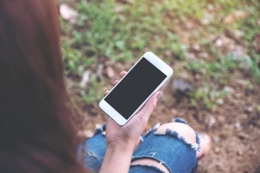 Yeşil bahçede otururken elinde boş siyah masaüstü ekran olan beyaz cep telefonu tutan kadının görüntüsü