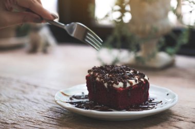 Çatal kullanan bir kadın, kırmızı kadife pastayı kremayla ve çikolatayla ahşap masada beyaz seramik tabakta kesiyor.