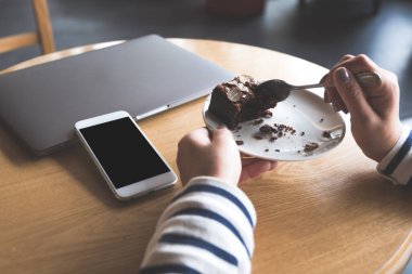 Laptopun yanında boş siyah masaüstü ekranlı beyaz bir cep telefonunun kek yiyen bir kadınla görüntüsü.