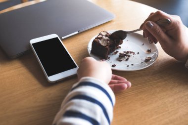 Laptopun yanında boş siyah masaüstü ekranlı beyaz bir cep telefonunun kek yiyen bir kadınla görüntüsü.