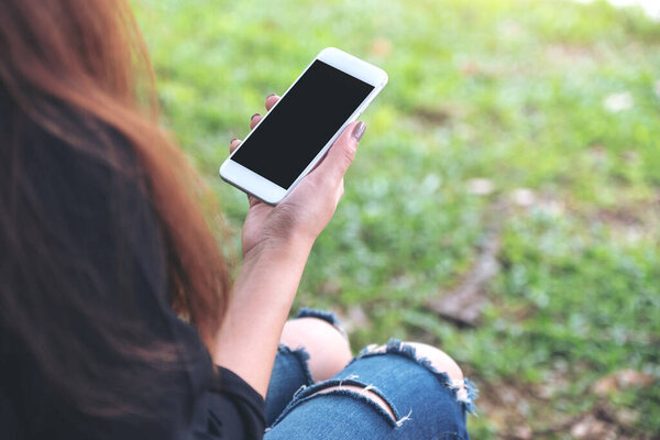 Mockup image of woman holding white mobile phone with blank black desktop screen while sitting on green yard