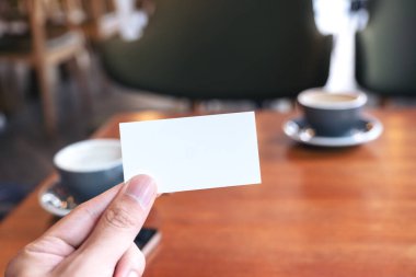 Kafedeki ahşap masada iki kahve fincanı olan beyaz, boş bir kartviziti tutan bir el.