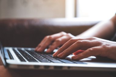 Ahşap masadaki, bulanık arkaplanlı dizüstü bilgisayarda çalışan ve yazı yazan ellerin yakın plan resmi