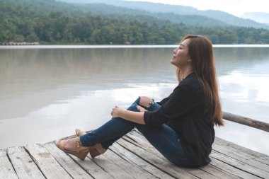 Nehrin kıyısındaki eski ahşap iskelede yalnız oturan Asyalı bir kadının görüntüsü. Mutlu, gökyüzü ve dağ arkası.