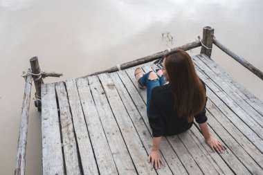 Asyalı bir kadının nehir kıyısındaki eski bir ahşap iskelede tek başına oturmasının en üst görüntüsü. 
