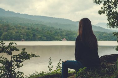 Gökyüzü ve dağ arkası ile nehir kenarında yalnız oturan Asyalı bir kadının yakın plan görüntüsü. 