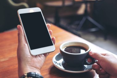 Modern kafede kahve içerken beyaz cep telefonlu, siyah ekranlı ellerin görüntüsü.