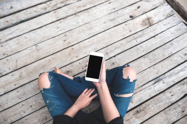 Üst görüntüde, eski ahşap zemin zemininde otururken elinde boş siyah ekranlı beyaz cep telefonu olan bir kadının görüntüsü var.