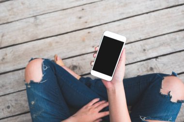 Üst görüntüde, eski ahşap zemin zemininde otururken elinde boş siyah ekranlı beyaz cep telefonu olan bir kadının görüntüsü var.