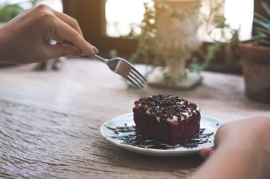 Çatal kullanan bir kadın, kırmızı kadife pastayı kremayla ve çikolatayla ahşap masada beyaz seramik tabakta kesiyor.