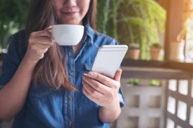 Bir kadının elinde tuttuğu, kahve içerken akıllı bir telefona dokunduğu yakın plan görüntüsü.