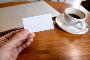 Ofisteki ahşap masada dizüstü bilgisayarı ve kahve fincanı olan beyaz, boş bir kartviziti tutan bir el.