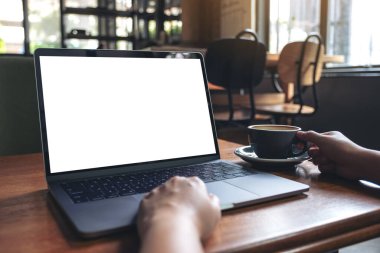 Kafedeki ahşap masada sıcak kahve içerken bilgisayarını beyaz ekranla kullanan bir kadının görüntüsü.