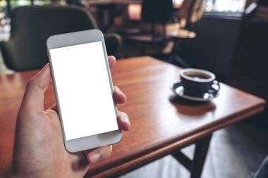 Kafedeki ahşap masada kahve fincanı ve boş masaüstü ekranı olan beyaz cep telefonlu el görüntüsü.