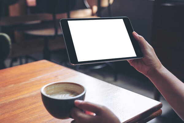 Образец женских рук, держащих черный планшет с чистым белым экраном с кофейной чашкой на деревянном столе в кафе
