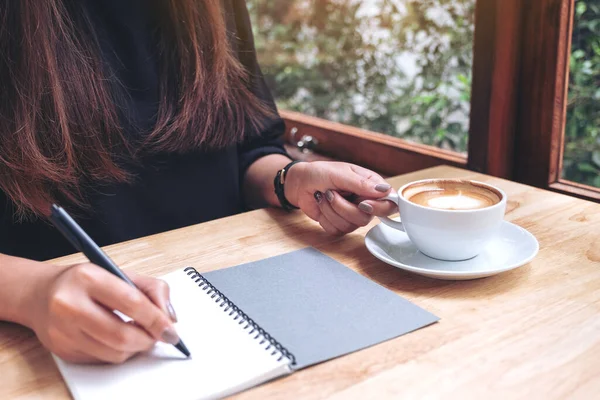 在木制桌子上喝咖啡时 一个女人的手在白色空白笔记本上写下的照片 — 图库照片