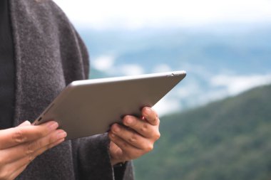 Mavi arkaplanlı sisli bir günde yeşil dağlı tablet tableti tutan ve kullanan bir kadının yakın plan görüntüsü