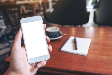 Kahve fincanı ve kafedeki ahşap masada defter ile boş masaüstü ekranı olan beyaz cep telefonlu el görüntüsü.