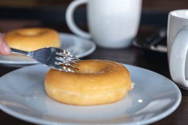 Masada kahve fincanları olan beyaz bir tabakta çatalla bir parça çörek kesen bir el.