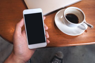 Üst görüntüde, kafedeki ahşap masada, kahve fincanı ve dizüstü bilgisayarlı, boş, siyah masaüstü ekranlı beyaz bir cep telefonu tutan el görüntüsü var.