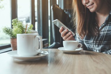 Kafede kahve fincanlarıyla akıllı bir telefonu tutan, kullanan ve dokunan Asyalı bir kadının yakın plan görüntüsü.