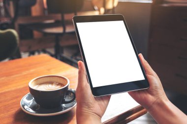 Kafe 'deki ahşap masada kahve fincanı ve beyaz ekranlı siyah tableti tutan ellerin görüntüsü.
