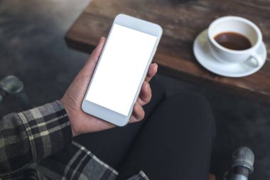 Kafedeki ahşap masada kahve fincanı ve boş masaüstü ekranı olan beyaz cep telefonlu bir kadının el resmi.