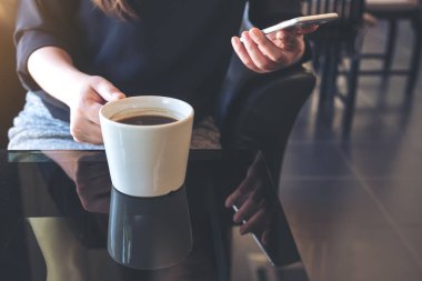 Kafede kahve içerken elinde telefon tutan ve kullanan bir kadının yakın plan görüntüsü.