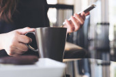 Detailní záběr ženy držící a používající smartphone při pití kávy v kavárně