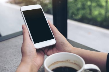 Kahve masasında kahve fincanı olan, boş siyah masaüstü ekranlı, beyaz cep telefonu tutan ellerin görüntüsü.